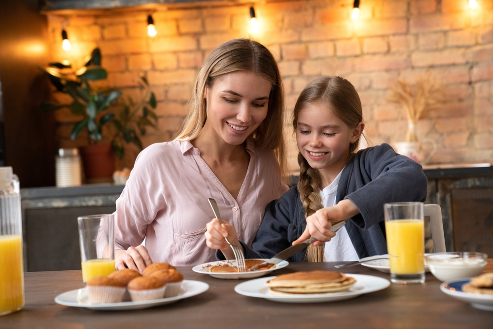 Une petite fille souriante partage un morceau de crêpe dans une assiette avec sa mère lors d'un brunch à l'occasion de la fête des mères dans une cuisine moderne. 