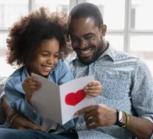 La petite fille a fait une surprise à son père, assis sur le canapé en lisant des mots agréables et chaleureux sur une carte qu'elle a fait pour la Fête des Pères, avec un grand coeur rouge en signe d'amour pour son père.