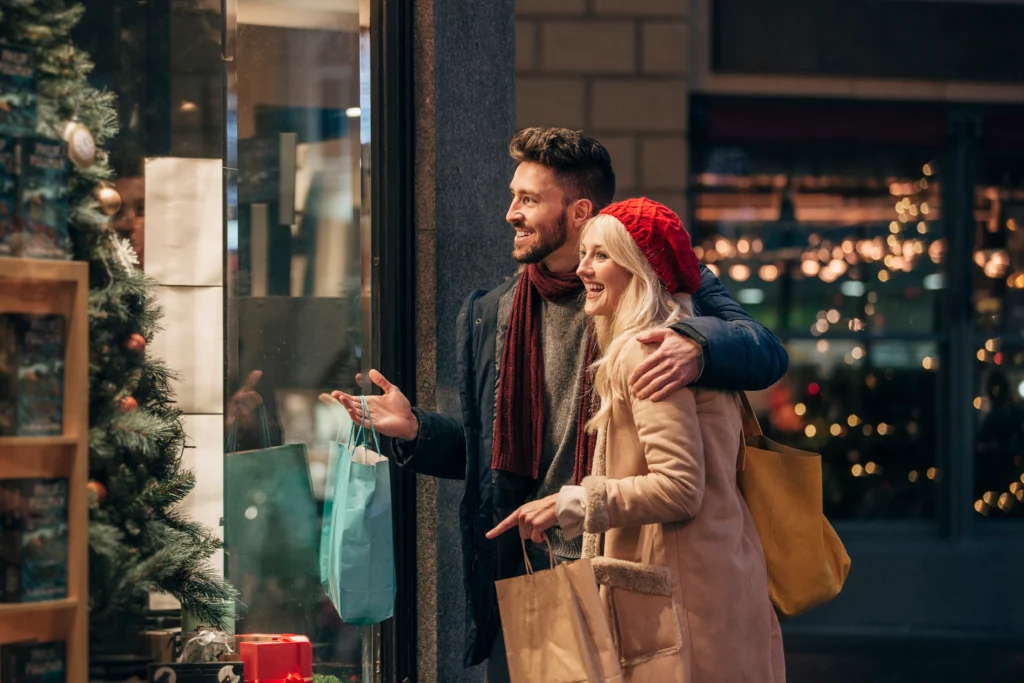 Vue de face d'un couple faisant des achats de noel pendant le black friday. L'homme adulte a son bras autour de la femme adulte et ils parlent de ce qu'il y a dans la vitrine.