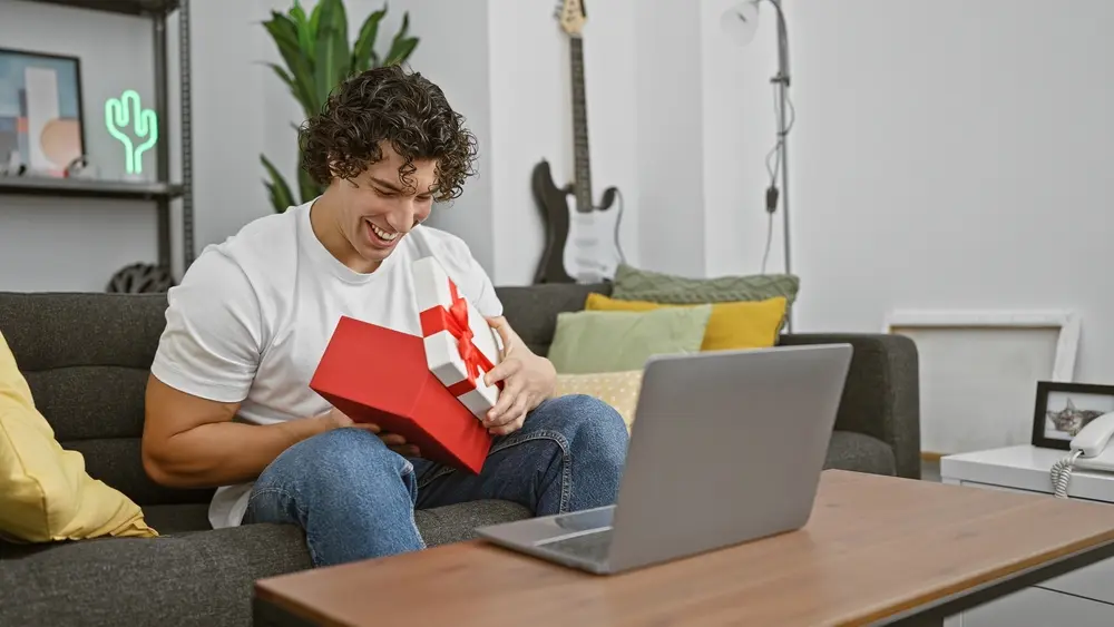 Un jeune homme souriant à la maison a ouvert avec enthousiasme un coffret cadeau Smartbox Black friday, avec un ordinateur portable sur la table indiquant une célébration virtuelle.