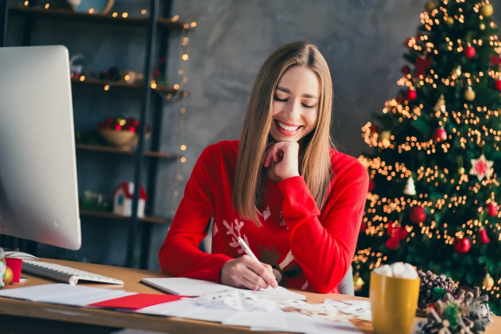 Photo d'une jeune femme qui rédige sa wishlist de noël sur un bureau, habillé en tenue de noël confortable.