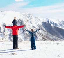 Un parent et deux enfants profitant d'une vue panoramique sur une chaîne de montagnes enneigée sous un ciel bleu clair. Le parent tient un enfant sur ses épaules, tandis que l'autre étend ses bras en émerveillement.