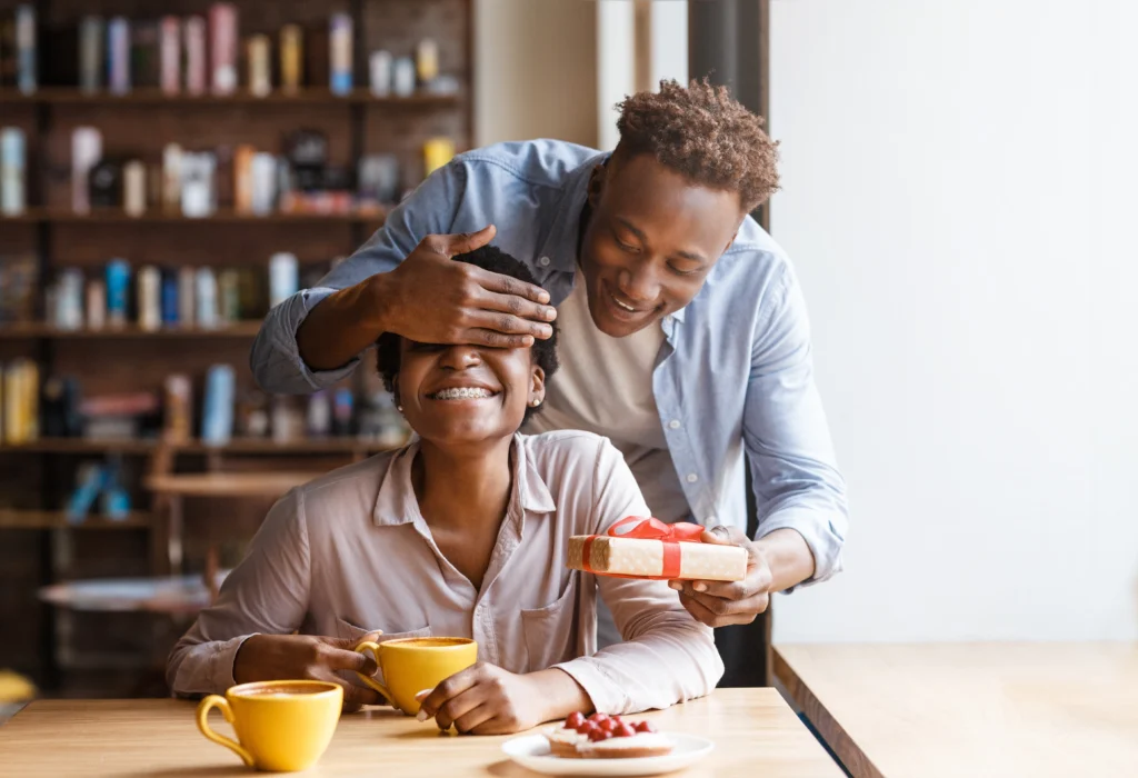 Joli jeune homme noir fermant les yeux de sa petite amie pour faire la surprise dans le café-restaurant urbain à l'occasion de la Saint-Valentin.