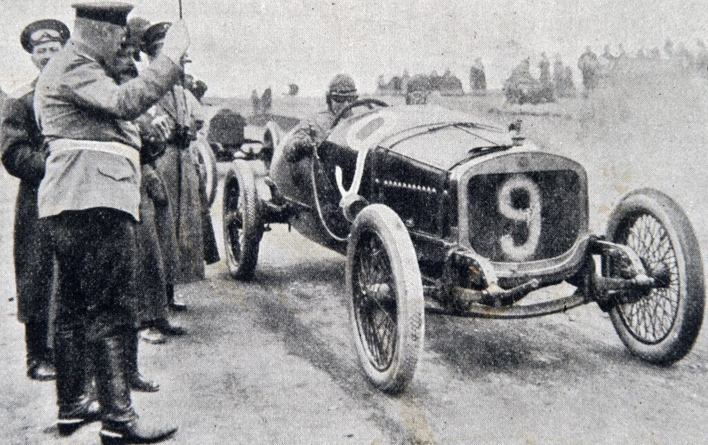 Foto antigua de piloto en un coche de carreras rodeado de gente